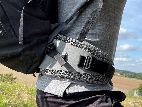 The waist of a man wearing a gray t-shirt with the lattice structure padding of the backpack tied around it. In the background a field, trees and blue sky with white clouds.